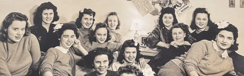 Een klassieke foto van een groepje jonge studentes in een kamer, allemaal lachend naar de camera, met versierde muur op de achtergrond.